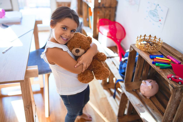 Очаровательный малыш улыбается счастливый обнимающий плюшевого мишку вокруг много игрушек в детском саду
 - Фото, изображение