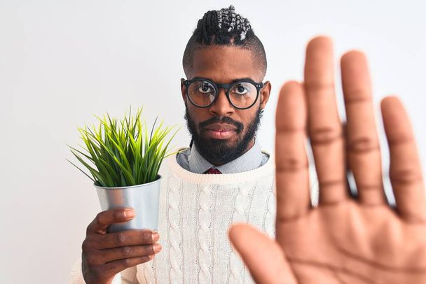 Homme afro-américain avec des tresses tenant pot de plante sur fond blanc isolé avec la main ouverte faisant stop signe avec expression sérieuse et confiante, geste de défense
 - Photo, image
