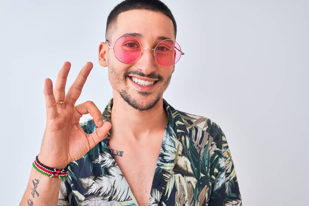 Молодой красивый мужчина в гавайской рубашке и розовых солнцезащитных очках на изолированном фоне делает хорошо знак с пальцами, отличный символ
 - Фото, изображение