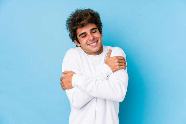Young caucasian man against a blue background isolated hugs, smiling carefree and happy. - Photo, Image