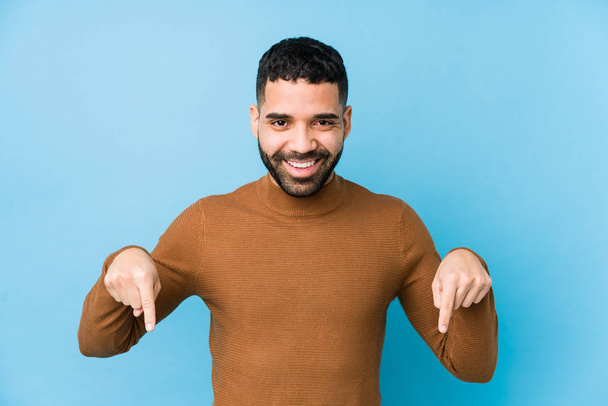 Homem latino jovem contra um fundo azul isolado aponta para baixo com os dedos, sensação positiva
. - Foto, Imagem