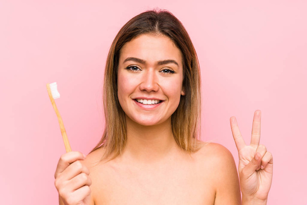 Νεαρή λευκή γυναίκα που κρατάει μια οδοντόβουρτσα απομονωμένη και δείχνει το νούμερο δύο με δάχτυλα.. - Φωτογραφία, εικόνα