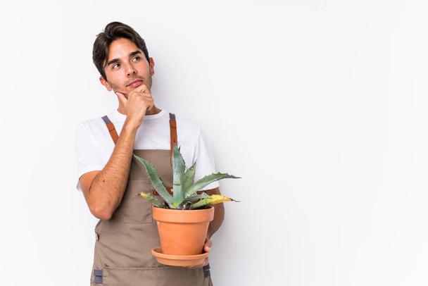 Homem jardineiro caucasiano jovem segurando uma planta isolada olhando para os lados com expressão duvidosa e cética
. - Foto, Imagem