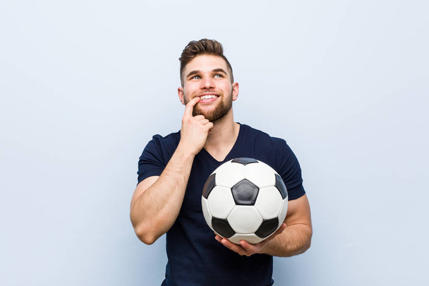 Młody biały mężczyzna trzymający piłkę nożną zrelaksowany myśląc o czymś patrząc na przestrzeń kopii. - Zdjęcie, obraz