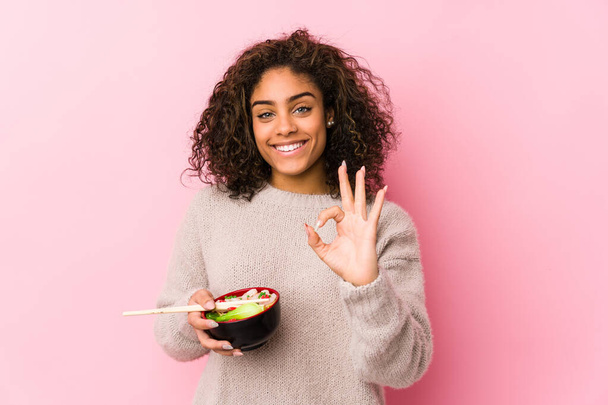 junge afrikanisch-amerikanische Frau essen Nudeln fröhlich und zuversichtlich zeigt ok Geste. - Foto, Bild