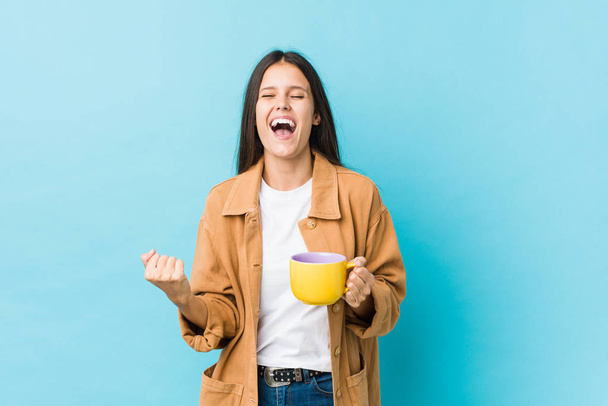 Kafkasyalı genç bir kadın kahve fincanı tutuyor tasasız ve heyecanlı bir şekilde tezahürat yapıyor. Zafer konsepti. - Fotoğraf, Görsel