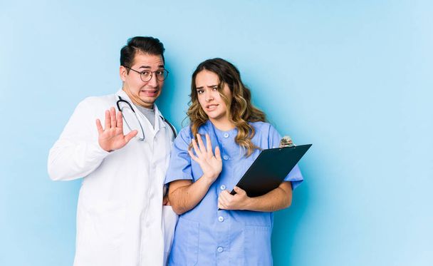 Joven pareja de médicos posando en un fondo azul aislado rechazando a alguien mostrando un gesto de disgusto
. - Foto, Imagen