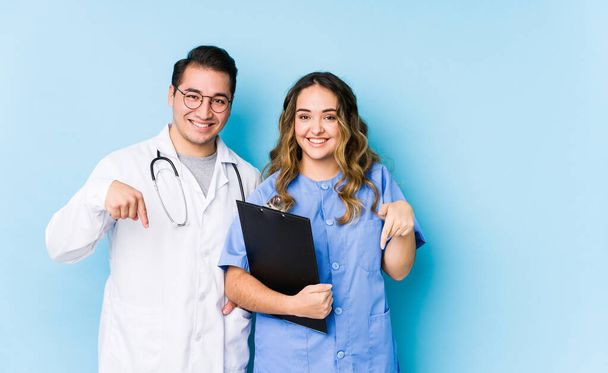 Jovem casal médico posando em um fundo azul isolado aponta para baixo com os dedos, sentimento positivo
. - Foto, Imagem