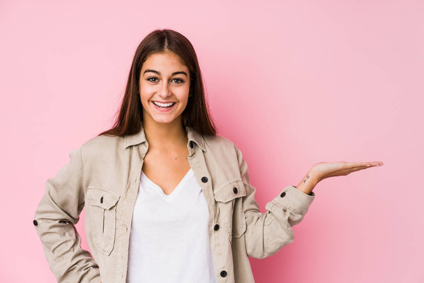 Young caucasian woman posing in a pink background showing a copy space on a palm and holding another hand on waist. - Photo, Image