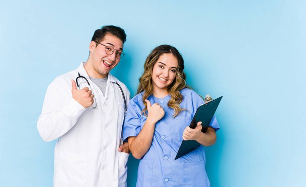 Молодая пара врачей позирует на синем фоне изолировано поднимая оба больших пальца вверх, улыбаясь и уверенно
. - Фото, изображение