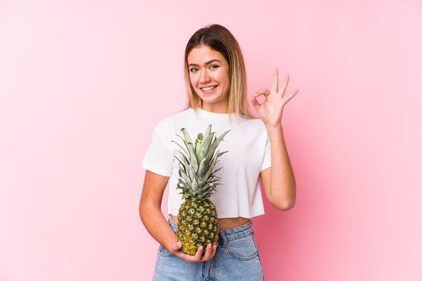 Молода біла жінка тримає ананас веселий і впевнений, показуючи нормальний жест
. - Фото, зображення