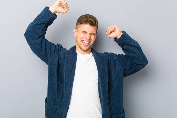 Fiatal kaukázusi férfi pizsamában ünnepel egy különleges napot, ugrik és felemeli a karját energiával. - Fotó, kép