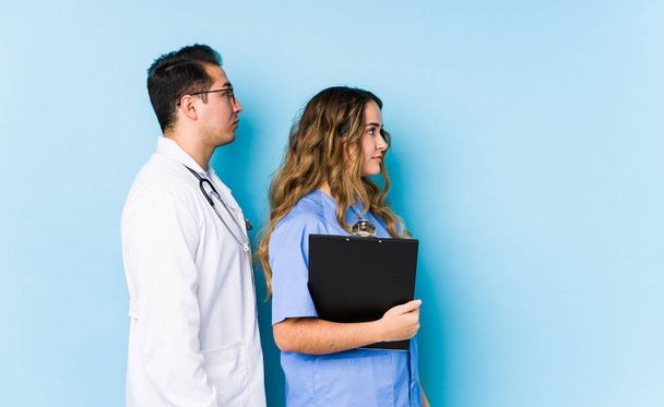 Joven pareja de médicos posando en un fondo azul mirando fijamente hacia la izquierda, de lado posan
. - Foto, Imagen