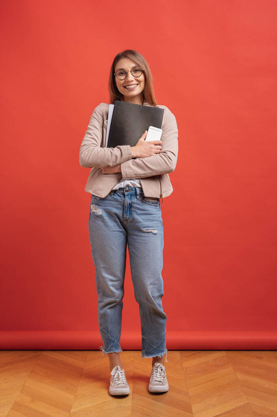Young smiling student or intern in eyeglasses standing with a folder on red background. - Foto, imagen