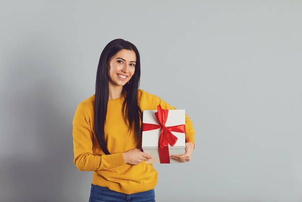 Happy girl with a gift box with a red ribbon on gray background. - Φωτογραφία, εικόνα