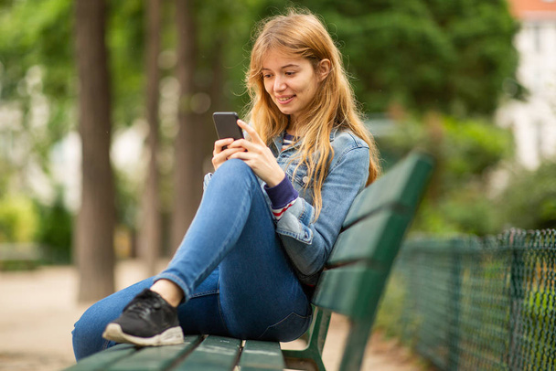 Porträt junge Frau sitzt auf Parkbank und schaut aufs Handy - Foto, Bild
