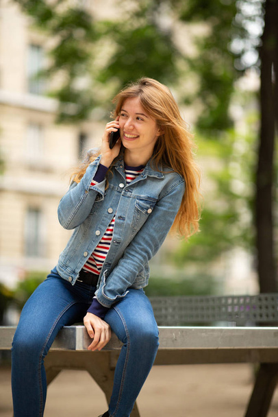 Retrato de uma jovem feliz sentada do lado de fora no parque conversando com celular
 - Foto, Imagem