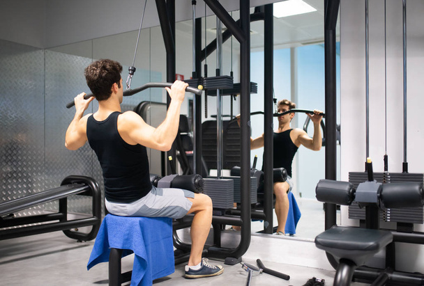 Hombre usando una máquina de lat en un gimnasio para entrenar sus hombros y espalda, culturismo y concepto de fitness natural - Foto, Imagen