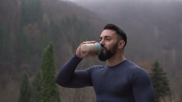 Hombre fuerte saludable con barba bebiendo Proteína Beber al aire libre con bosque brumoso en el fondo
 - Metraje, vídeo