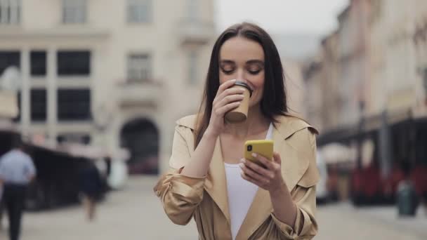 Young Pretty Lady Holding Coffe Cup Smiling Using her Smartphone Walking at City Street Close Up. - Séquence, vidéo