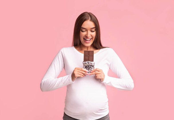 Беременная женщина ест шоколад на фоне розовой студии
 - Фото, изображение