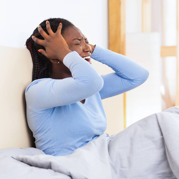 infeliz afro-americano senhora gritando tocando cabeça sentado na cama
 - Foto, Imagem
