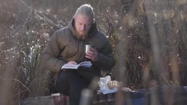 Thoughtful Caucasian man sitting on tree trunk, reading and drinking hot tea from steel mug. Lonely male tourist enjoying resting outdoors. Leisure, lifestyle, hobby. - Video