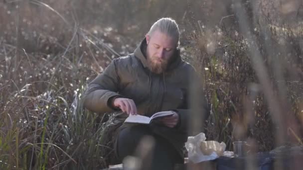 Portrait of thoughtful Caucasian man sitting on tree trunk in autumn forest and reading. Male tourist enjoying vacations outdoors. Leisure, lifestyle, hobby. - Záběry, video