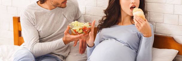 Mulher grávida rejeitando salada de legumes frescos e comer croissant
 - Foto, Imagem