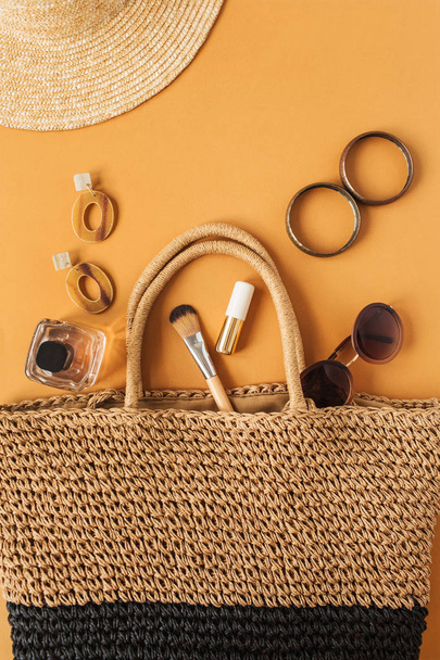 Hasır şapka, turuncu arka planda kadın aksesuarları olan rattan plaj çantası. Düz yerde, üst tarafta. Modern moda günlüğü. - Fotoğraf, Görsel
