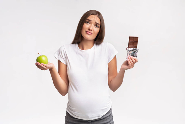 Confus femme enceinte tenant pomme et chocolat debout en studio
 - Photo, image