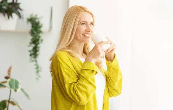 Onnellinen nainen pitelee teekuppia ja katselee ikkunaa
 - Valokuva, kuva