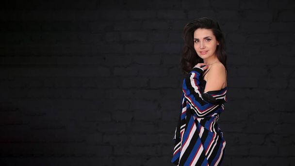 portrait of a beautiful young woman with dark hair in a black bodysuit and a striped cardigan in over the knee boots on a dark background - Zdjęcie, obraz