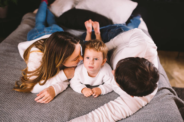 Happy family having fun lying down on bed at home. Happy Family In Bed. Mom dad and son lie on bedroom at within doors. father, mother and child relaxing bedstead in loft style apartment in evening - 写真・画像
