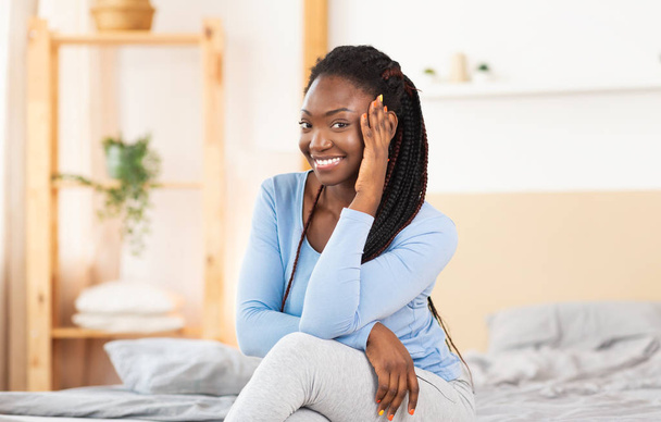 Mujer negra alegre posando sentada en una cómoda cama interior
 - Foto, Imagen
