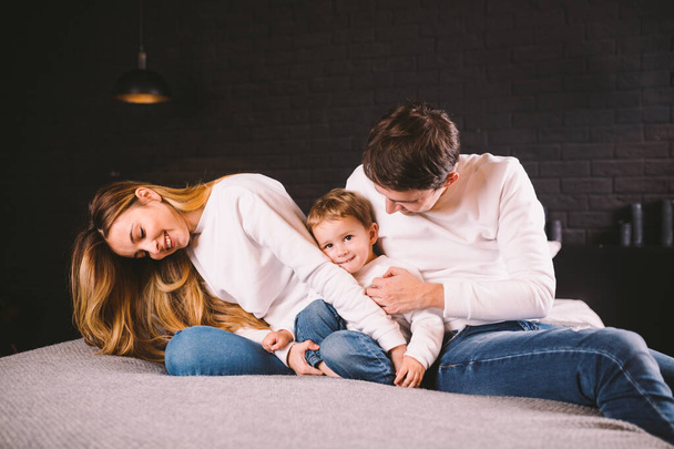 Parents with their baby boy on bed at home. Families with child in bed at night at home. Happy family enjoying in bed. Mom, dad and kid play in the bedroom in the evening. Loft style interior - Foto, imagen