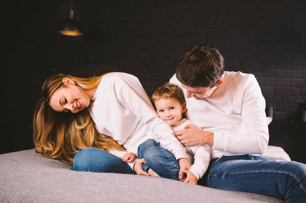 Happy family having fun lying down on bed at home. Happy Family In Bed. Mom dad and son lie on bedroom at within doors. father, mother and child relaxing bedstead in loft style apartment in evening - Foto, imagen