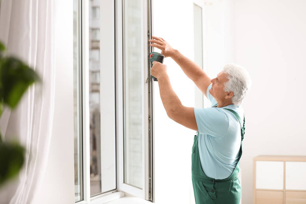 Зрелый строитель ремонтирует пластиковые окна с электрической отверткой в помещении
 - Фото, изображение