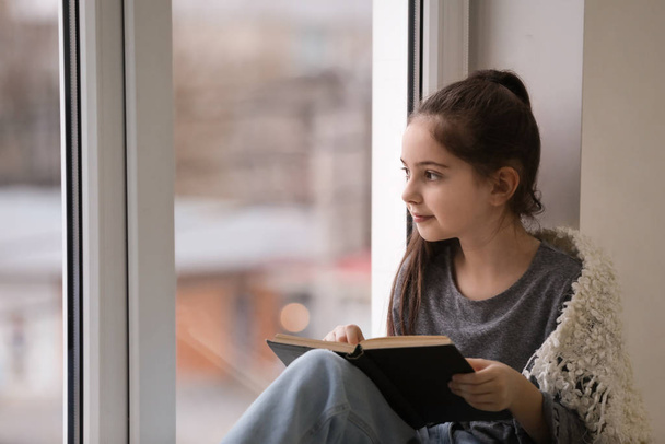 Cute little girl reading book near window at home. Space for text - Фото, изображение