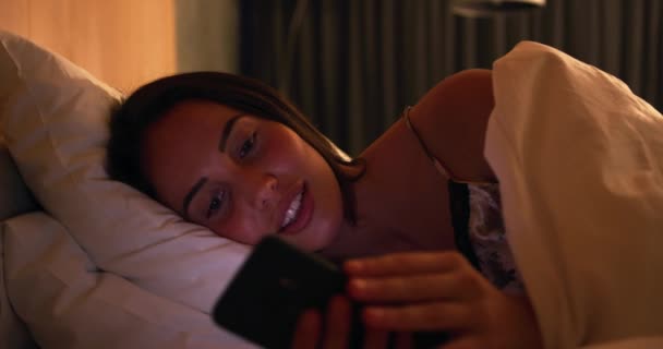 Front view of happy young Caucasian woman enjoying time off, lying in bed at night and using smartphone, slow motion - Кадры, видео
