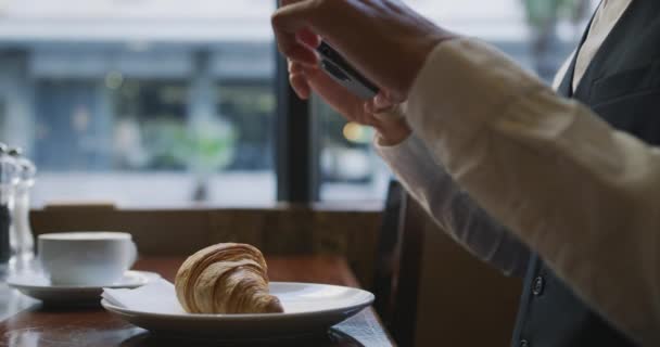 Side view of a young Caucasian elegant businessman in a cafe, taking photos of croissant with his smartphone, slow motion - Imágenes, Vídeo