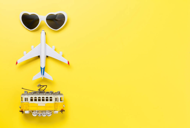 黄色の日当たりの良い背景にサングラス、トラム、飛行機のおもちゃで旅行のコンセプト。コピースペース付きのトップビューフラットレイ - 写真・画像