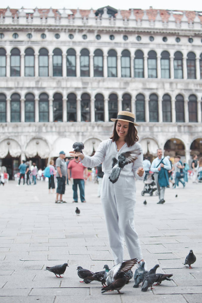 γυναίκα με λευκά ρούχα και ψάθινο καπέλο διασκεδάζει με περιστέρια στην πλατεία Venice City - Φωτογραφία, εικόνα