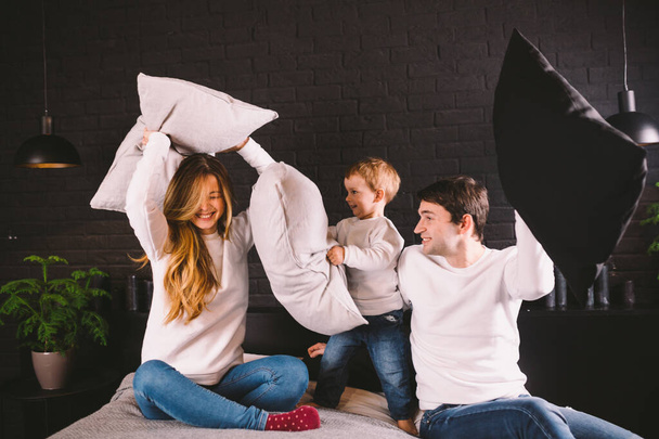 Family having funny pillow fight on bed. Parents spending free time with their son. Young family being playful at home. Happy parents and child. Young caucasian family fighting with pillows - Фото, изображение