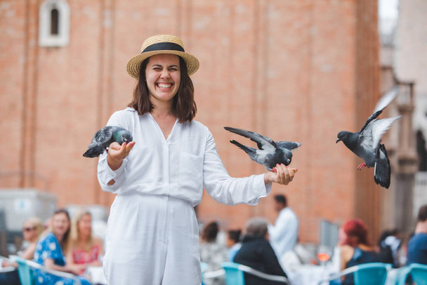 woman in white clothes with straw hat having fun with pigeons at venice city square - Photo, Image