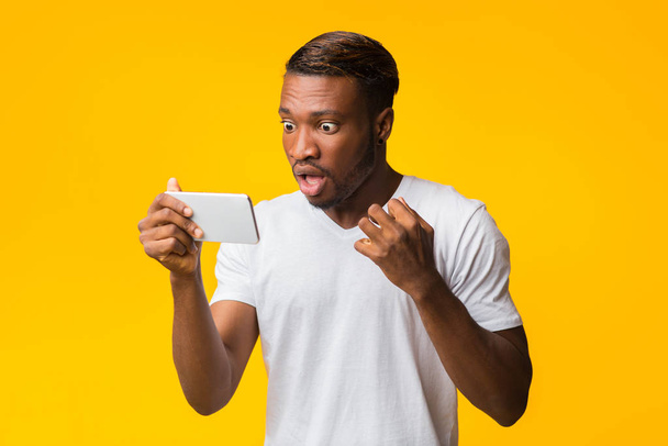 Geschokt Afro Guy op zoek naar Smartphone Staande in Studio - Foto, afbeelding