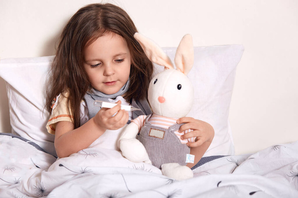 Little girl lying in bed and measuring soft hear's temperature with thermometer, child lying in bed on pillow with her favourite toy, wearing gray scarf around neck, suffering from flue, cathing cold. - Photo, Image