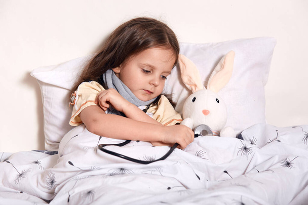 Portrait of caring sweet kid with scarf around neck lying in her bed with favourite toy, examining it with stethoscope, looking attentively at it, playing games alone. Children times concept. - Fotografie, Obrázek