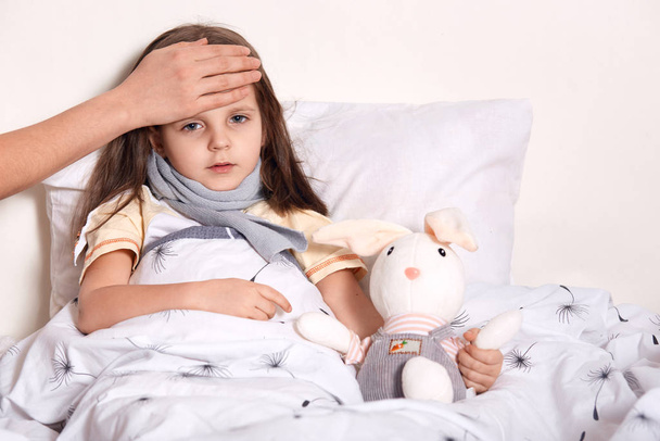 Indoor shot of little girl with fair hair lying in her bed, hugging favourite toy, having unknown hand on forehead, checking temperature, having scarf around neck, being on sick leave. Health concept. - Foto, Bild