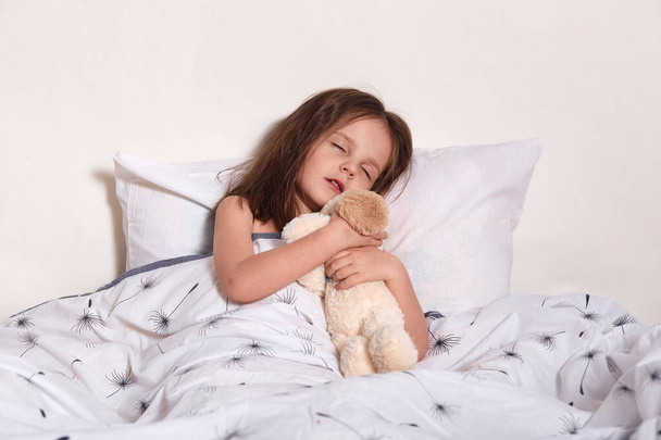 Image of charming magnetic little infant sleeping in her light bedroom alone, lying in soft cosy bed, hugging her favourite bear toy, closing eyes and opening mouth. Happy childhood concept. - Photo, image
